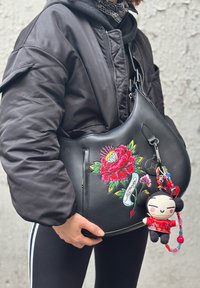 Melna ādas plecu soma ar košu ziedu izšuvumu un piekaramu mīksto taustiņu piekariņu, kas piesaistīts ar krāsainām pērlītēm.