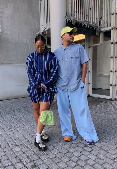 Dos personas están de pie sobre adoquines. Una lleva una camisa de rayas amplia en azul marino y pantalones cortos a juego, con gafas de sol verdes y una bolsa verde texturizada. La otra lleva una camisa abotonada azul claro, pantalones sueltos azules y zapatillas de colores.