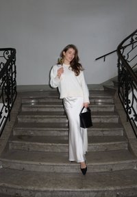 Λευκό πουλόβερ, σατέν ασημί φούστα, μαύρη τσάντα, κρατώντας ένα ποτήρι σαμπάνιας, στέκεται σε μάρμαρο σκαλοπάτια με στολισμένη μαύρη κουπαστή στο φόντο.