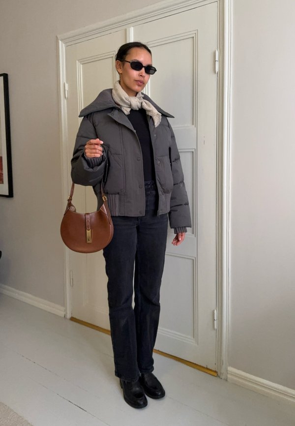 Femme portant une doudoune grise, un pantalon noir, des chaussures noires, une écharpe beige et des lunettes de soleil, tenant un sac à main marron à l'intérieur près d'une porte blanche.