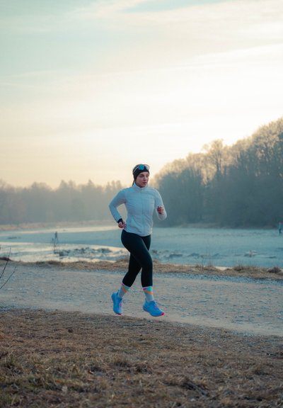 Mujer con chaqueta clara y leggins negros corriendo por un sendero junto a un río, con árboles y un cielo nublado al fondo.