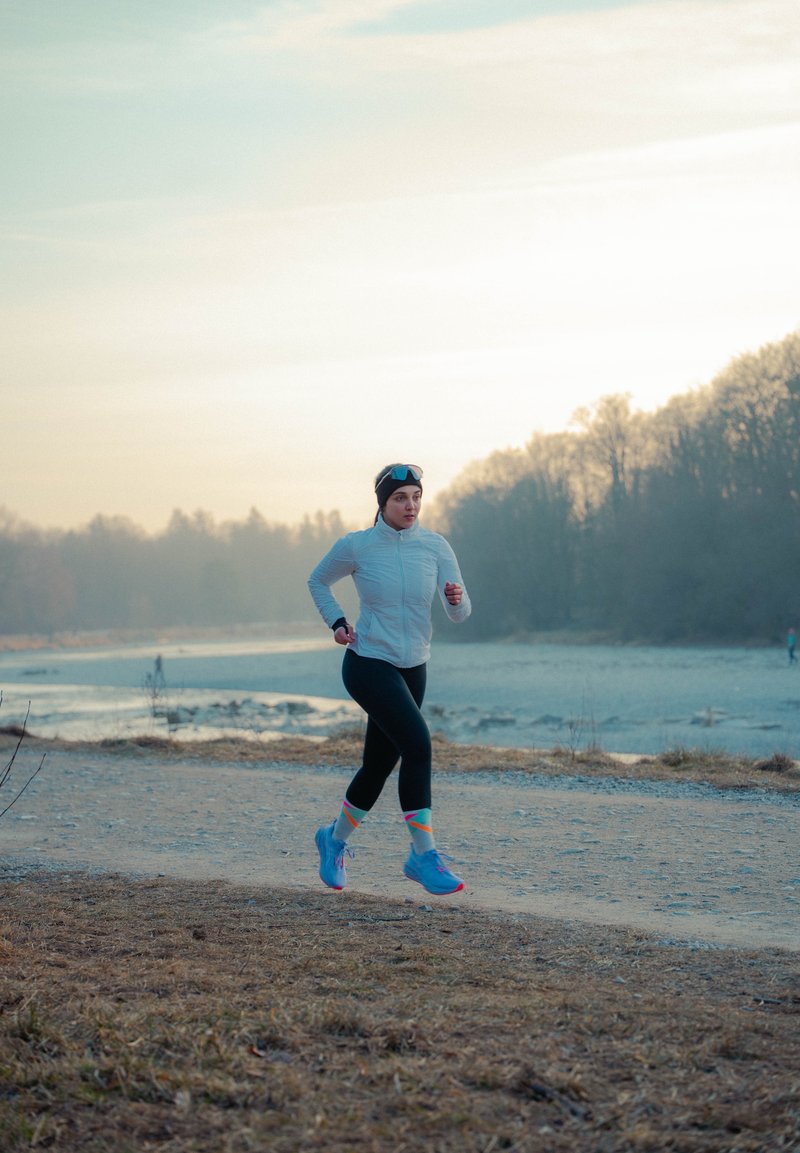 Femme en veste légère et leggings noirs courant sur un sentier près d'une rivière, avec des arbres et un ciel couvert en arrière-plan.