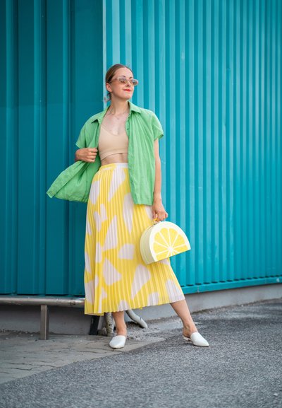 Camisa verde de manga corta, top corto beige, falda amarilla plisada con patrón abstracto blanco y un bolso en forma de limón. Zapatillas blancas sin cordones.