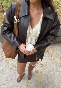 Barna bőrdzseki ezüst szív brossal, fehér csipkés felső, barna miniszoknya, barna térdcsizma, kávéscsészét tartva.