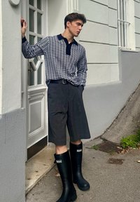 Crno-bijela prugasta polo majica dugih rukava, sivi karirani šorts i crne gumene čizme za kišu. Ova odjeća ima kontrastni ovratnik i pojas oko struka.