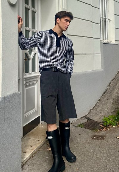 Polo de manga larga a rayas blanco y negro, pantalones cortos de cuadros grises y botas de lluvia de goma negras. El conjunto cuenta con un cuello a contraste y un cinturón en la cintura.