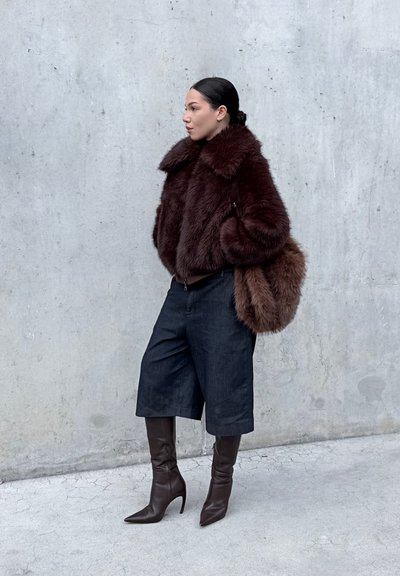 Abrigo marrón de piel sintética con gran cuello, shorts de mezclilla oscura y botas marrones con tacón que llegan hasta la rodilla. Bolso de hombro marrón y textura lisa.