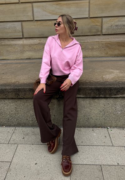 Sudadera rosa claro con cremallera, pantalones anchos marrones y zapatos marrones con cordones y suela gruesa. Los accesorios incluyen gafas de sol redondas negras.