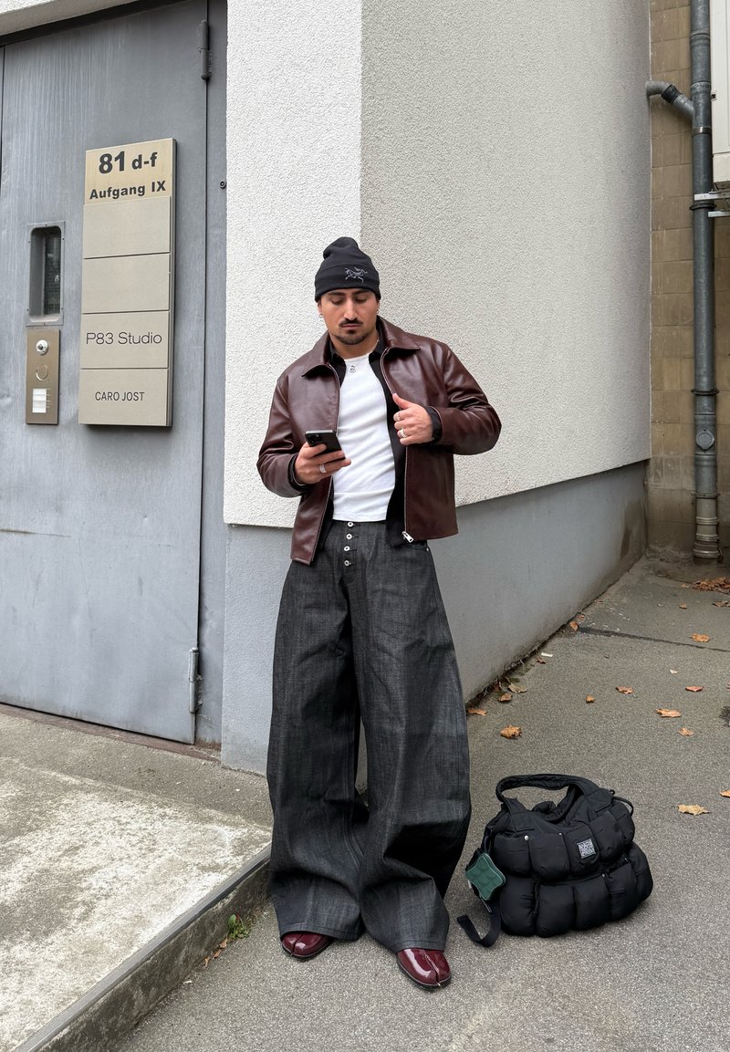 Homme portant une veste en cuir marron, un pantalon large foncé, un bonnet noir et des chaussures bordeaux, se tient près de l'entrée d'un bâtiment gris, utilisant un smartphone, avec un sac noir à proximité.