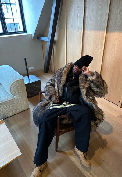 Hombre con un gran abrigo de piel y un gorro negro encorvado en una silla de madera en una habitación moderna con suelo y muebles de madera clara.