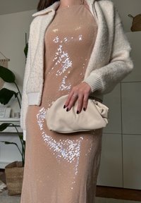 Γυναίκα με μπεζ φόρεμα με παγιέτες και λευκό χνουδωτό σακάκι, κρατώντας ένα κρεμ τσαντάκι φάκελο, με σκουρόχρωμο βερνίκι νυχιών, σε δωμάτιο με ξύλινο πάτωμα.