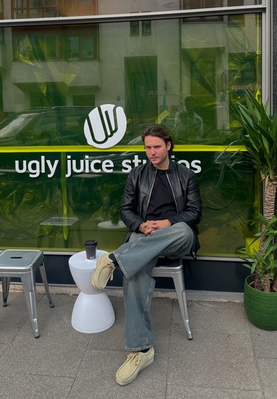 Chaqueta de cuero negra, camisa negra, jeans acampanados de color azul claro, zapatos beiges. Sentado cerca de un taburete blanco y una planta verde, con una taza de café.