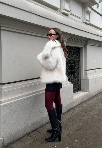 Mujer con abrigo blanco de piel sintética, gafas de sol negras, falda negra, medias burdeos y botas negras con tacón, de pie en la acera junto a una verja de metal ornamental.