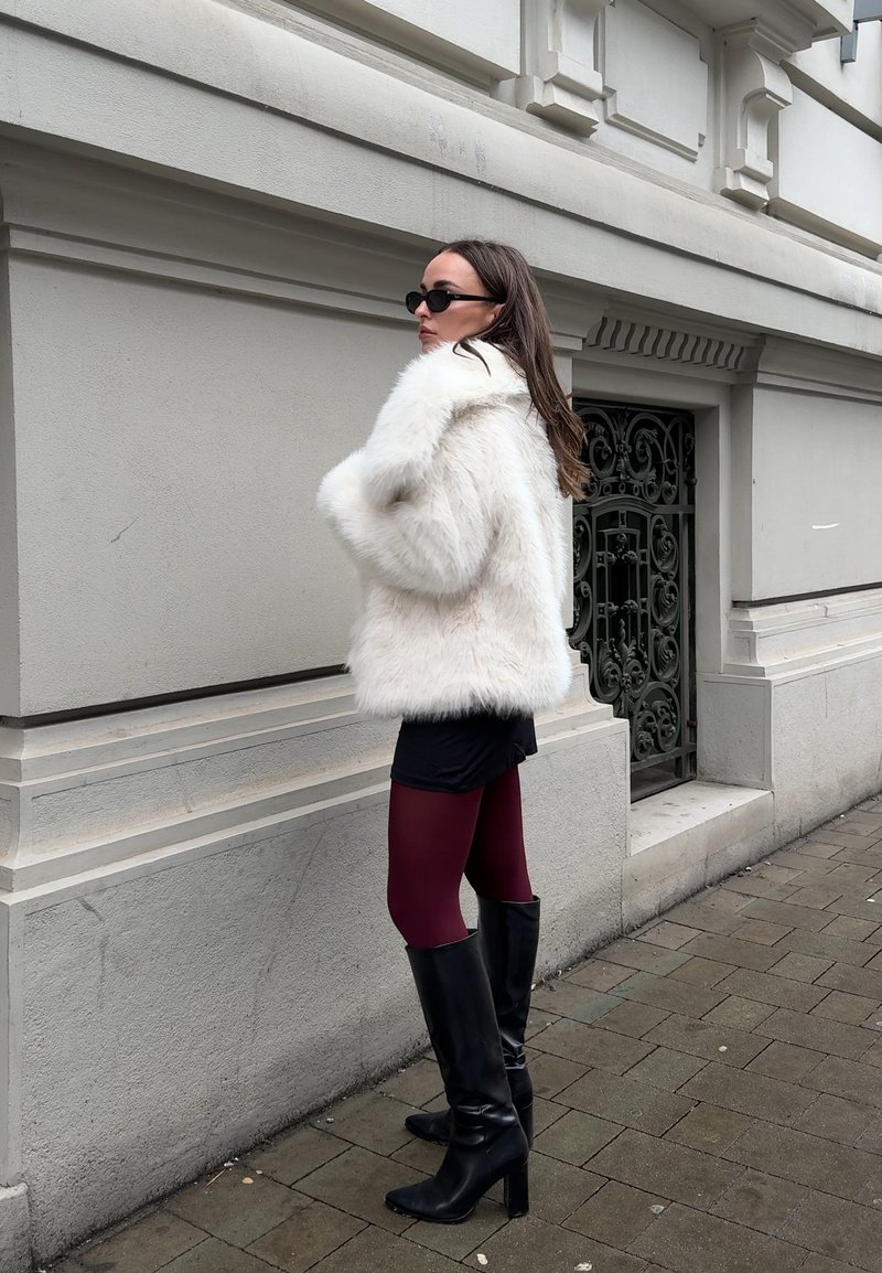 Donna con cappotto in pelliccia sintetica bianca, occhiali da sole neri, gonna nera, collant bordeaux e stivali neri con tacco in piedi sul marciapiede accanto a un cancello in metallo decorato.