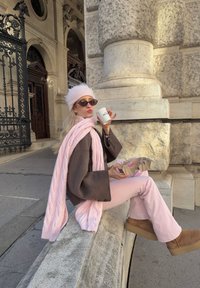 Hnědý oversize kabát, růžový pletený šátek, světle růžové kalhoty, béžové boty. Drží hrnek s kávou a světle hnědou papírovou krabičku na jídlo.