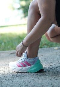 Bílé sportovní boty s růžovými detaily a mintově zelenou podrážkou, s lehkou konstrukcí a texturovaným svrškem, je zavazována osobou.