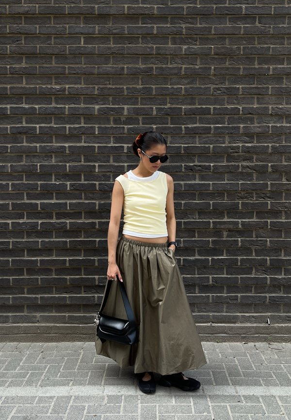 Jeune femme portant des lunettes de soleil et un haut jaune sans manches, tenant un sac à main noir, debout contre un mur de briques sombre.