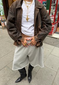 Casaco oversized castanho, camisa curta branca, calções cinzentos e botas pretas de cano alto com bico fino. Colares e anéis em camadas douradas. Estilo urbano casual.