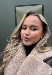 Jeune femme aux longs cheveux blonds portant un manteau en shearling beige et un col roulé noir, posant avec confiance sur un fond vert.