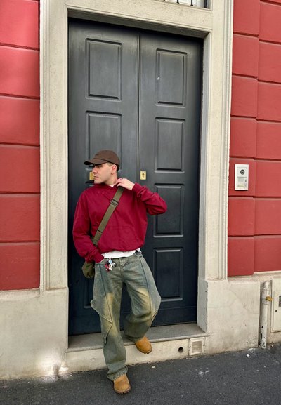 Sudadera roja, camiseta blanca, vaqueros verdes holgados, gorra marrón y botas beige. Apoyado contra una puerta oscura con un marco blanco y pared roja.