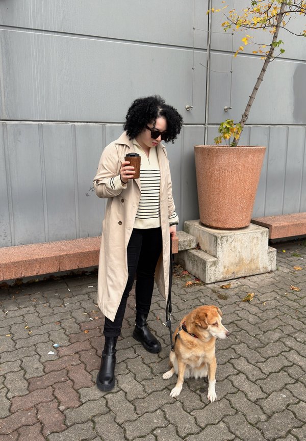 Cappotto beige, maglione a righe nere, pantaloni neri e stivali neri. Un cane è seduto vicino. Tazza di caffè in mano su uno sfondo grigio.