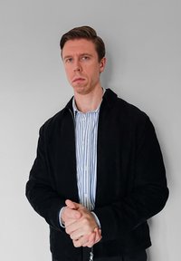 Veste noire zippée portée sur une chemise rayée bleu et blanc. Le modèle a les cheveux courts, les mains jointes et se tient devant un mur gris clair.