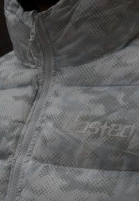 Polstret jakke i et lyst grått kamuflasjemønster, med høy krage, glidelås og preget logo på brystet i matchende farge.