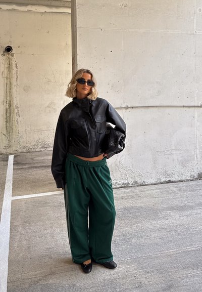 Mujer con cabello rubio corto, vestida con chaqueta de cuero negra, gafas de sol oscuras, pantalones anchos verdes, zapatos negros, sosteniendo un bolso tejido negro, de pie junto a una pared de concreto.