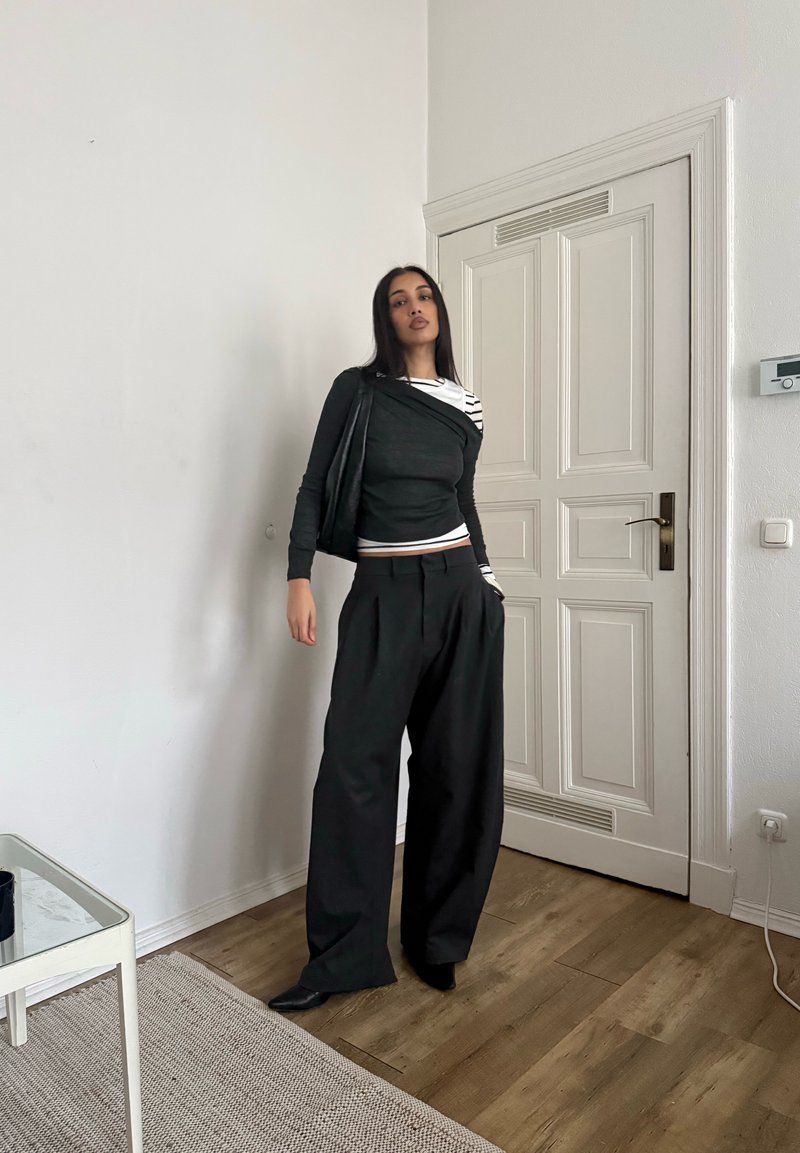 Femme debout à l'intérieur portant un pantalon noir large, un haut rayé noir et blanc asymétrique, avec une main dans la poche et un bras détendu.