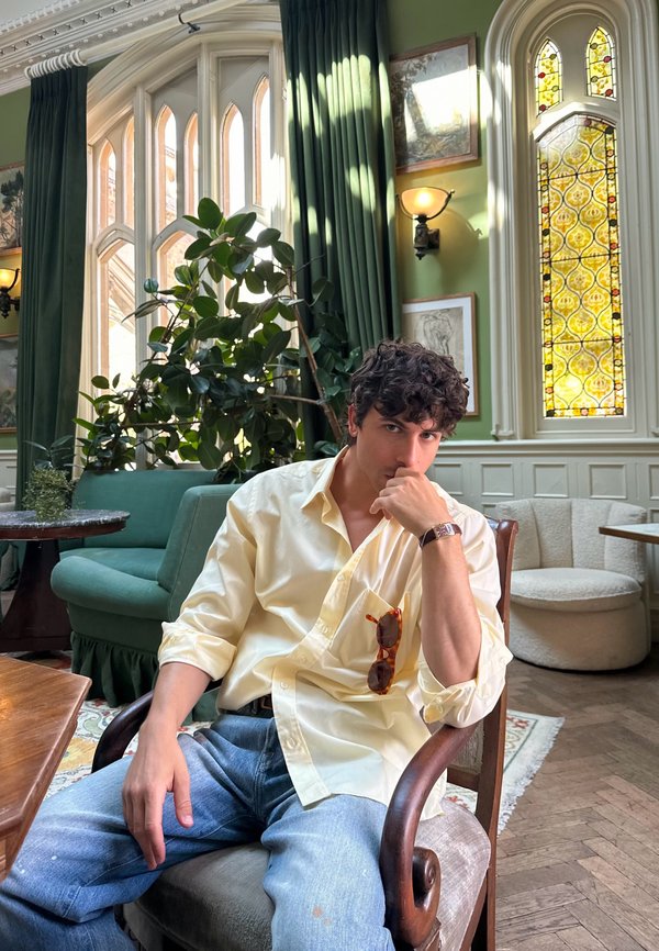 Jeune homme en chemise jaune et jean assis dans une pièce vintage avec des rideaux verts, une fenêtre en vitrail, des plantes et des meubles tapissés.