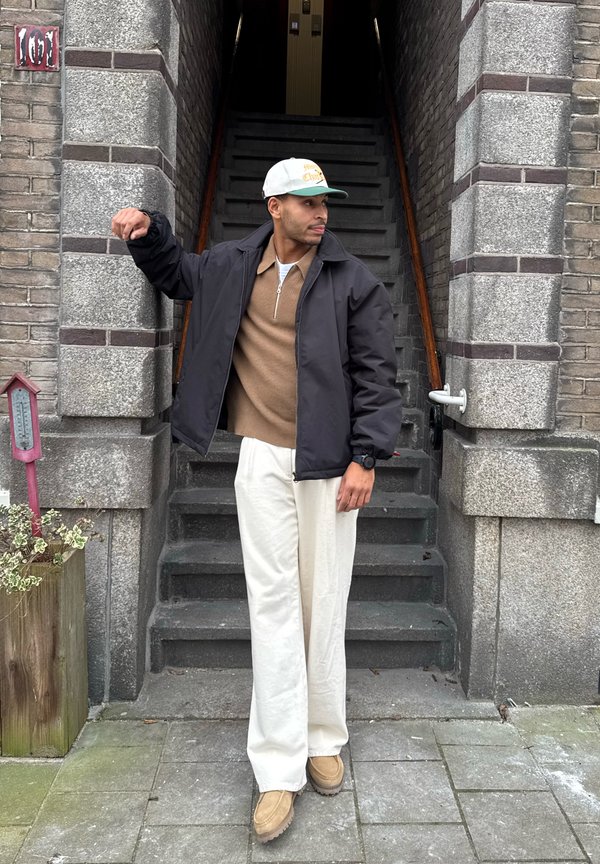 Vyras, vilkintis smėlio spalvos kelnes, rudą megztinį, juodą striukę ir baltą beisbolo kepuraitę, stovi prie akmeninių laiptų įėjimo, žiūrėdamas į kairę ir pakėlęs vieną ranką.