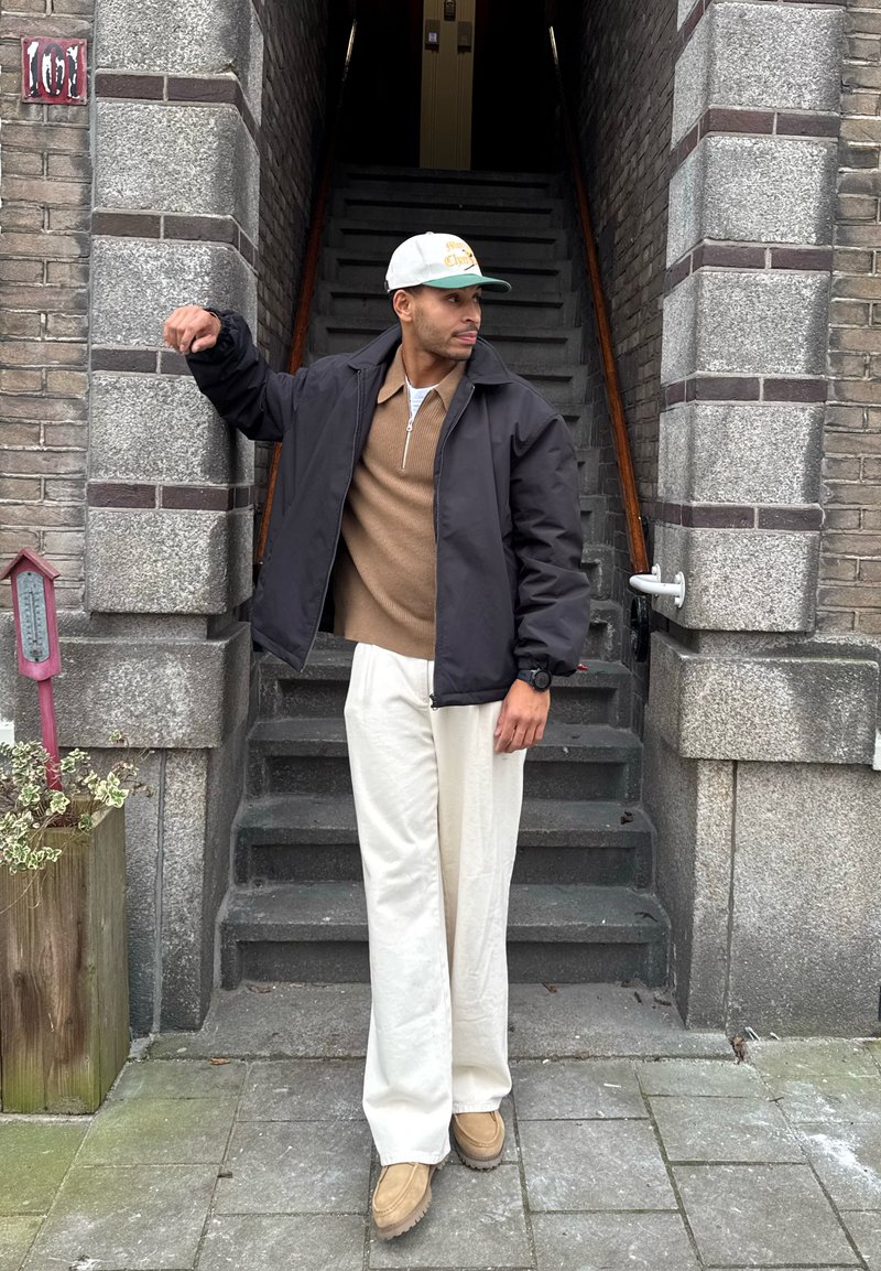 Homme portant un pantalon beige, un pull marron, une veste noire et une casquette de baseball blanche, debout près de l'entrée d'un escalier en pierre, regardant à gauche avec un bras levé.