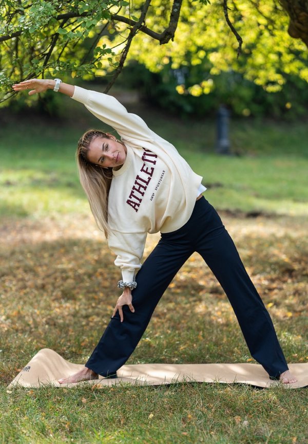Une femme s’étire latéralement sur un tapis sous des arbres feuillus dans un parc ensoleillé.