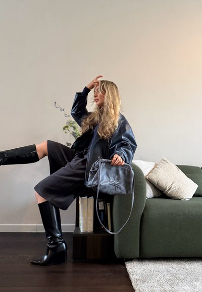 Mujer con botas negras, chaqueta de cuero negra y vestido oscuro se sienta en el brazo de un sofá verde, sosteniendo un bolso gris con libros y flores a su lado.