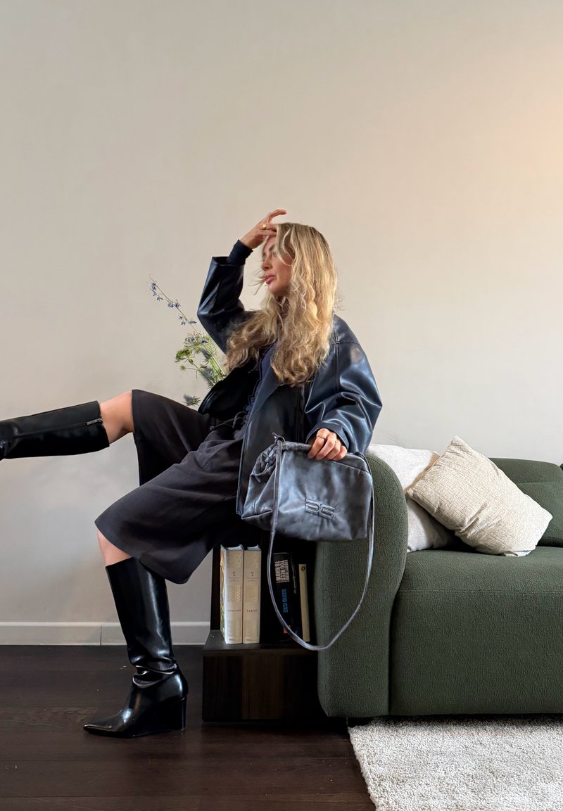 Femme portant des bottes noires, une veste en cuir noire et une robe foncée, assise sur l'accoudoir d'un canapé vert, tenant un sac à main gris, avec des livres et des fleurs à côté d'elle.