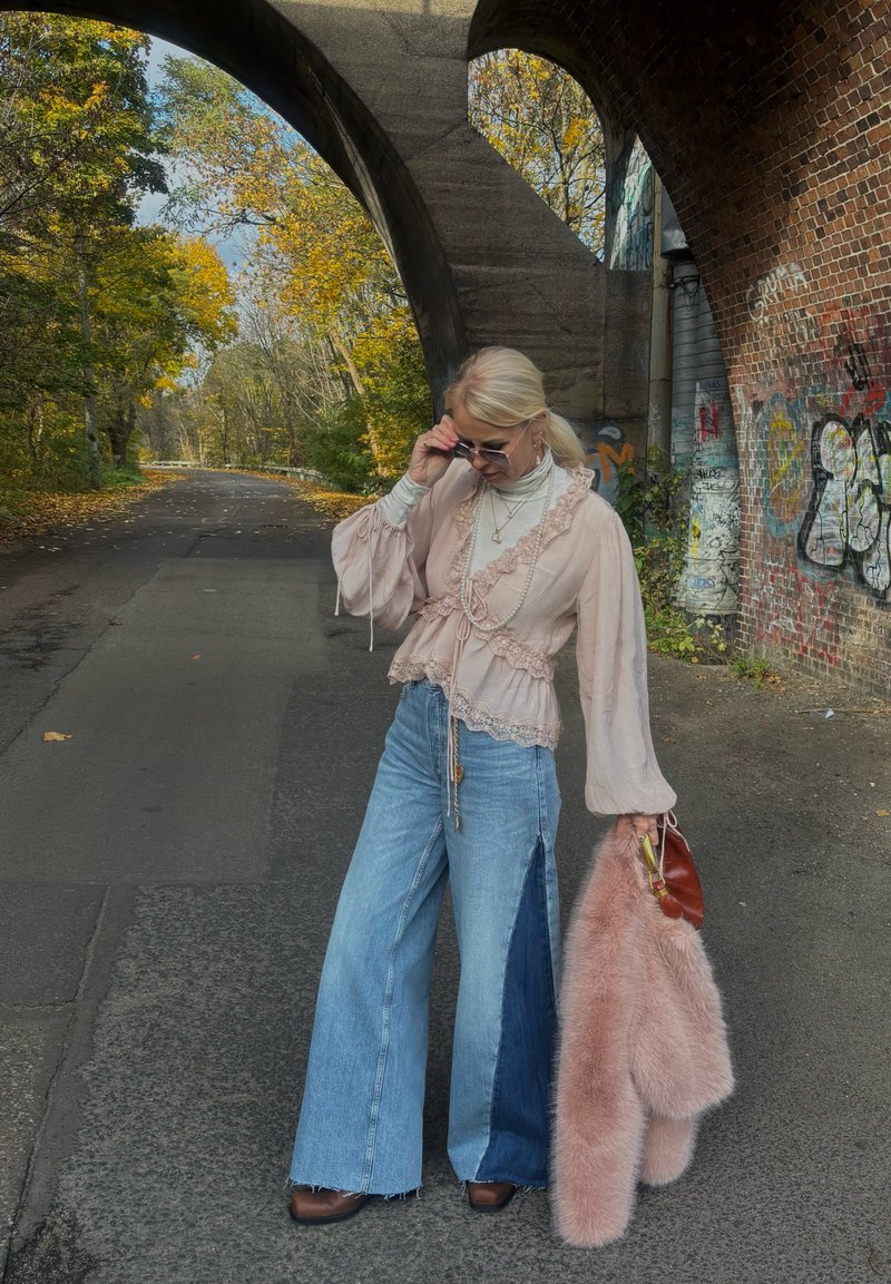 Light pink textured blouse with ruffled trim, paired with wide-leg blue jeans featuring a side panel. Holding a pink fur coat and handbag.