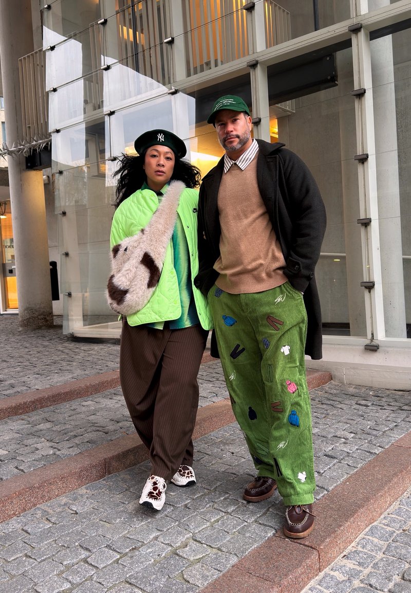 Deux personnes marchent sur des marches pavées devant un bâtiment moderne, portant des tenues colorées à motifs et des chapeaux.