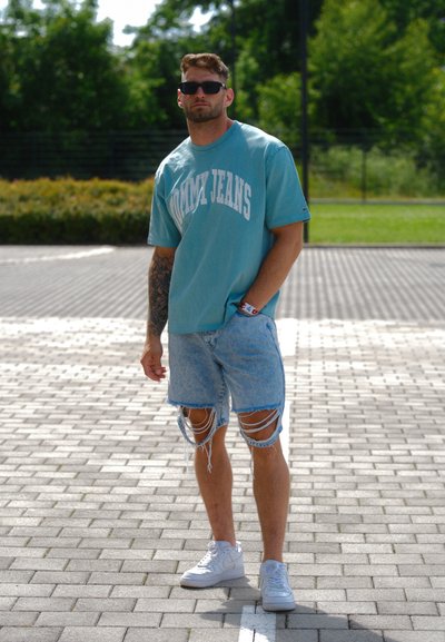 Camiseta de algodón de gran tamaño en azul claro con el texto "TOMMY JEANS", combinada con pantalones cortos de denim desgastados. Se lleva con zapatillas blancas y gafas de sol.