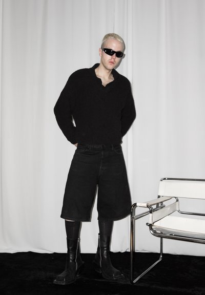 Persona con el cabello rubio corto que lleva gafas de sol negras, un suéter negro oversize, pantalones cortos anchos negros, medias y botas negras, de pie junto a una silla moderna.