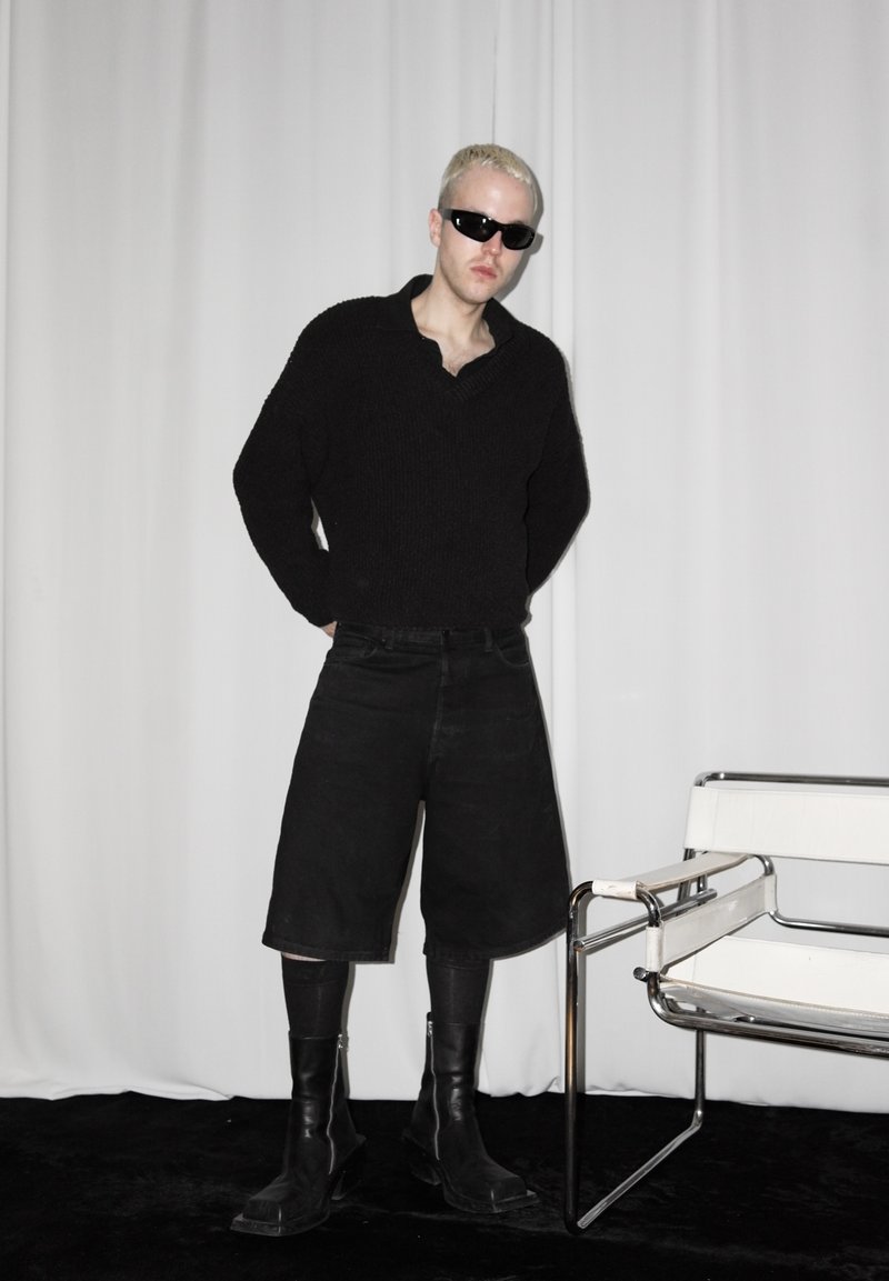 Personne aux cheveux blonds courts portant des lunettes de soleil noires, un pull noir surdimensionné, un short large noir, des collants et des bottes noires, debout à côté d'une chaise moderne.