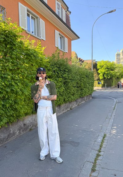 Hombre con tatuajes, gafas de sol, gorra, camiseta verde y pantalones blancos comiendo helado en la acera cerca de un edificio naranja y un seto verde.
