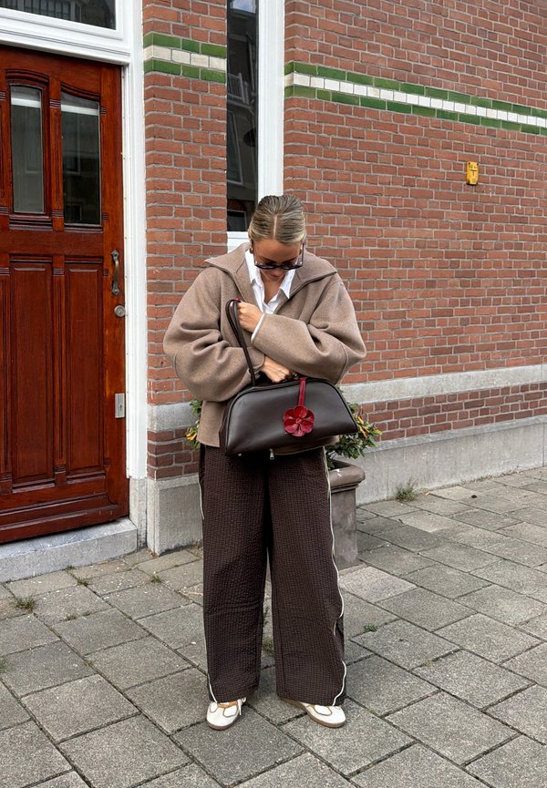 Femme portant une veste beige oversize et un pantalon marron large, tenant un sac marron foncé avec un porte-clés fleur rouge, debout sur un trottoir près d'un mur en briques.