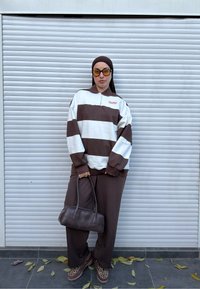 Henkilö, jolla on päällään ruskeavalkoraidallinen neulepaita, ruskeat housut, huivi, aurinkolasit ja joka pitää kädessään ruskeaa käsilaukkua, seisoo valkoisen säleikön edessä.