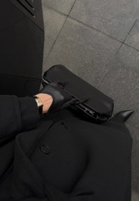 Svart skinnveske med glidelåslukking og to håndtak. En hånd med svart hanske holder vesken, kombinert med en frakk og spisse svarte sko.