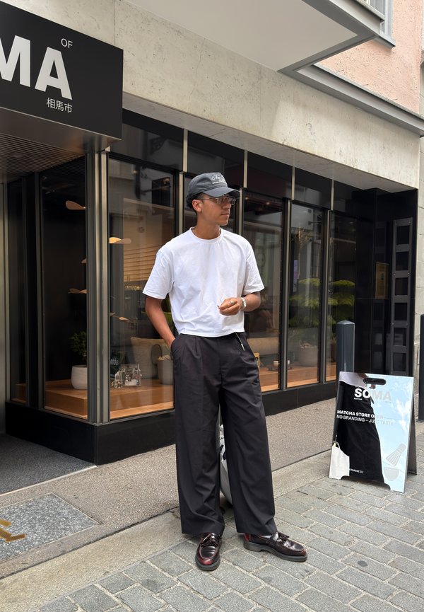 Mann trägt weißes T-Shirt, schwarze weite Hose, Kappe und Brille, steht auf dem Bürgersteig in der Stadt in der Nähe des Ladeneingangs und eines stehenden Schildes.