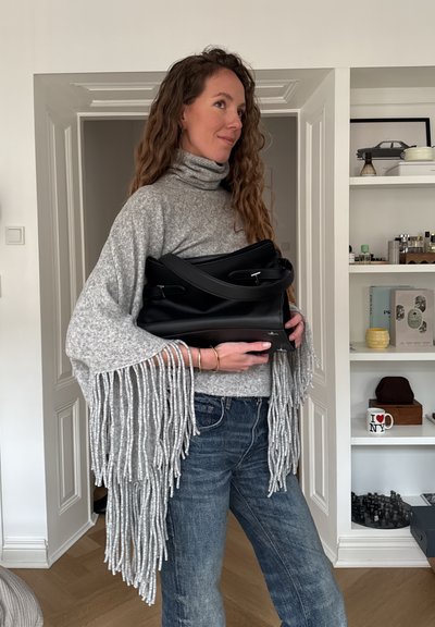 Mujer con cabello largo y rizado, vistiendo un suéter gris con flecos y jeans, sosteniendo un bolso grande negro en el interior cerca de un estante blanco.