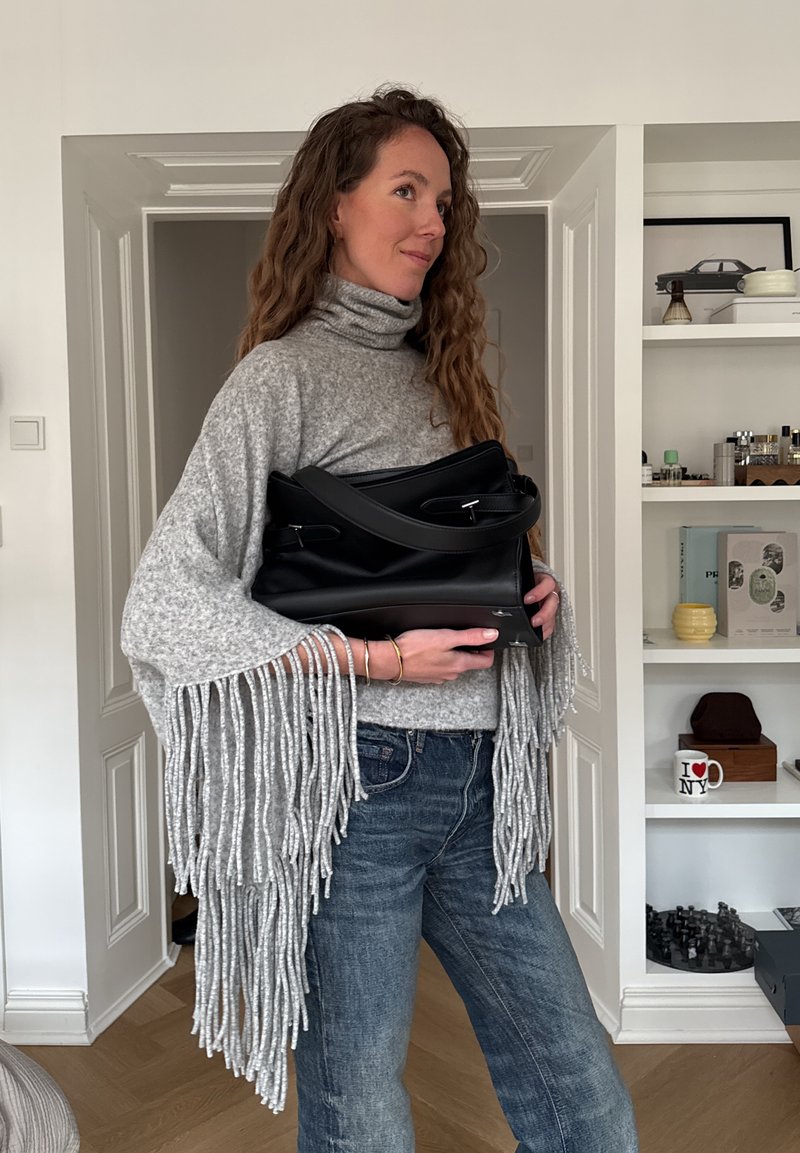 Femme aux longs cheveux bouclés portant un pull gris à franges et un jean, tenant un grand sac à main noir à l'intérieur près d'une étagère blanche.