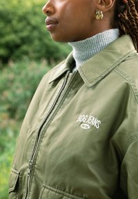 Olivově zelená bomber bunda se zipem, zdobená bílou výšivkou "BOG JEANS". Nositá přes světle šedý rolák. Texturovaná látka.