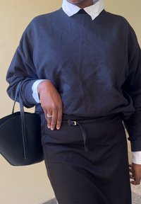 Schwarzer Baumwoll-oversized Sweatshirt, das über einem weißen Kragenhemd getragen wird, kombiniert mit einem schwarzen Rock. Hand ruht auf einer schwarzen Tote-Tasche.