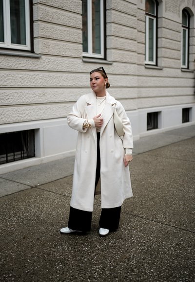 Abrigo blanco largo, pantalones de pierna ancha negros, zapatillas blancas. Accesorios mínimos que incluyen un bolso de color claro y joyería sutil. Superficie texturada.